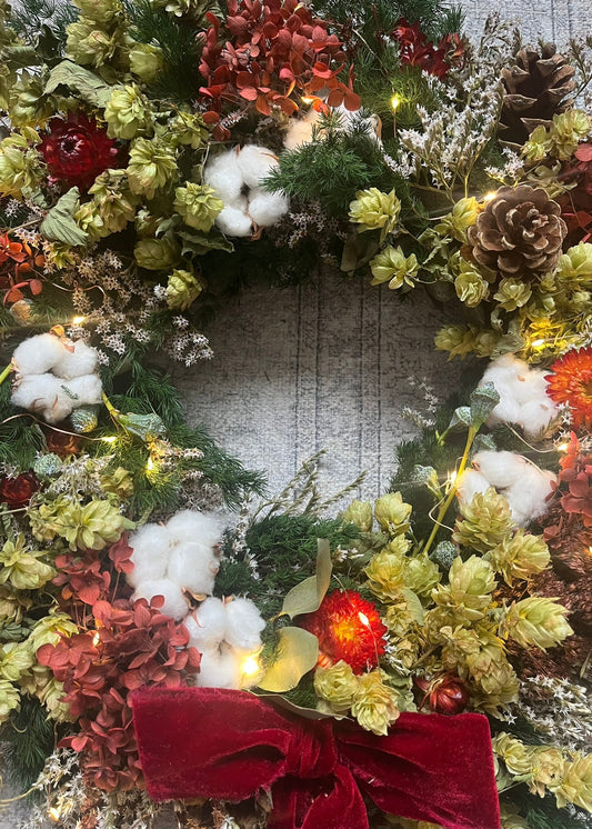 Decorative wreath with flowers, cotton, and a red bow on a textured surface.