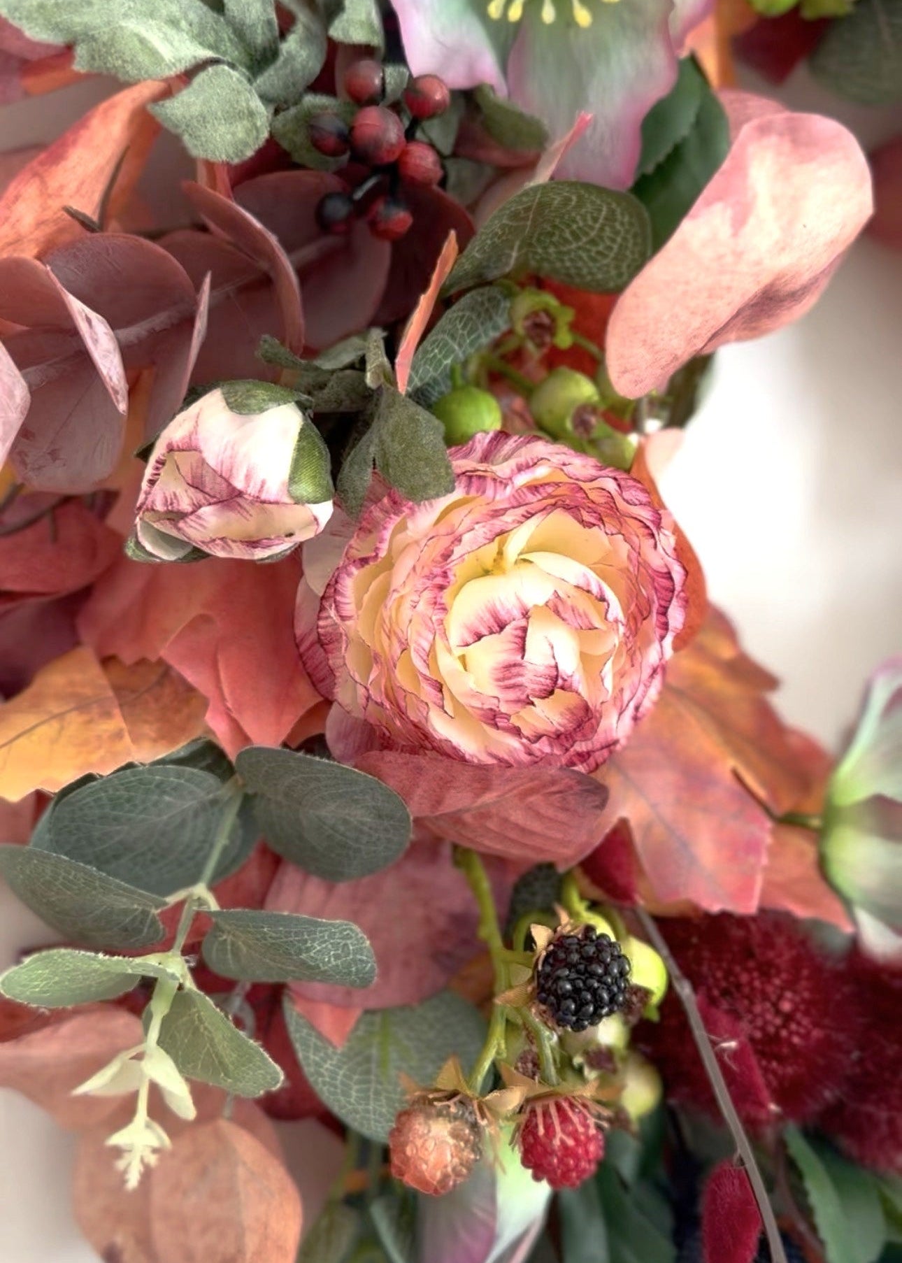 A close-up of an autumn floral arrangement featuring a central pink and cream ranunculus-like flower, dusty pink eucalyptus leaves, green hollyhocks, dark berries, and muted foliage.