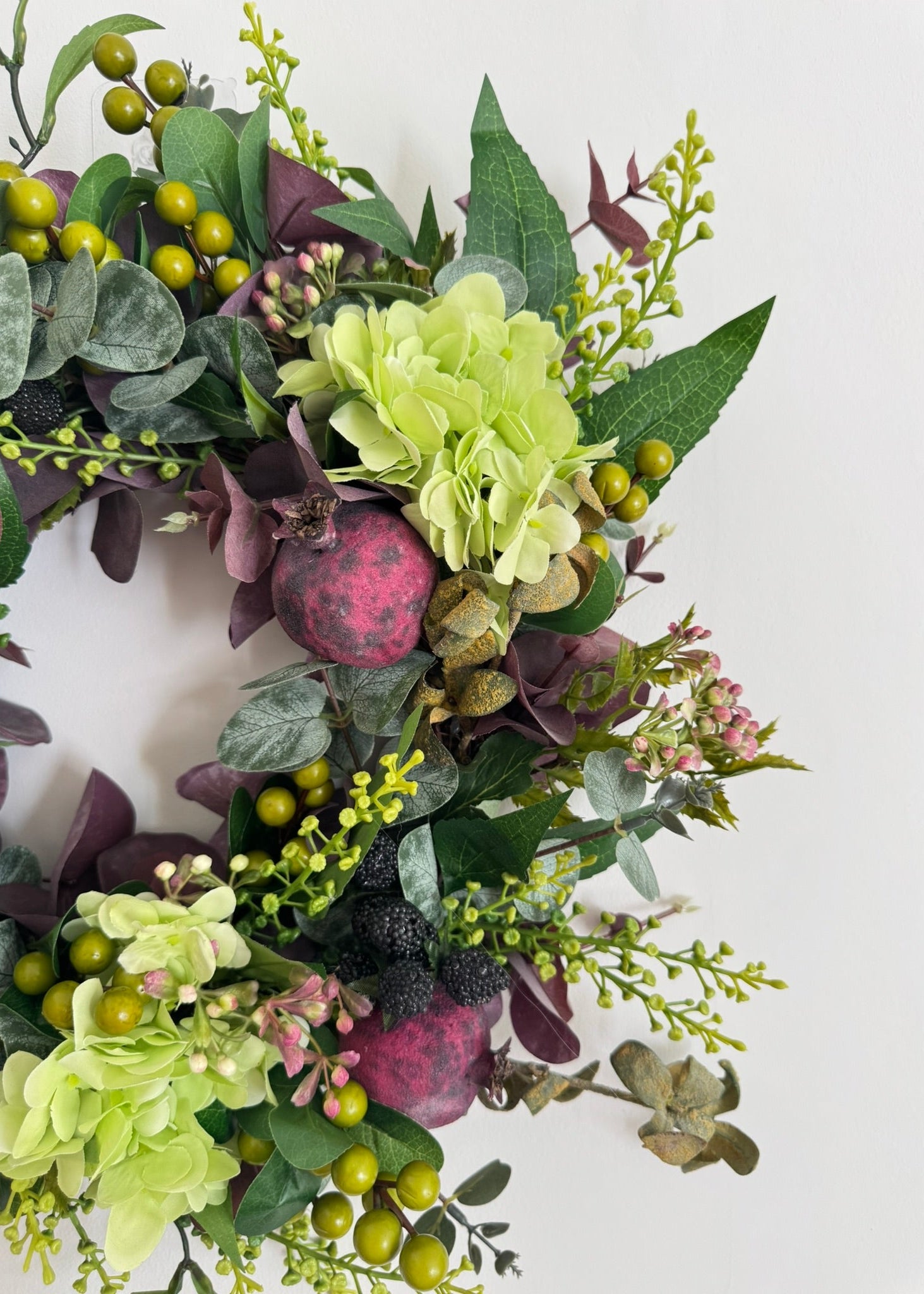 An artificial floral wreath featuring light green hydrangea-like blooms, dark green leaves, burgundy berries, and olive-green berries, arranged on a plain white background.