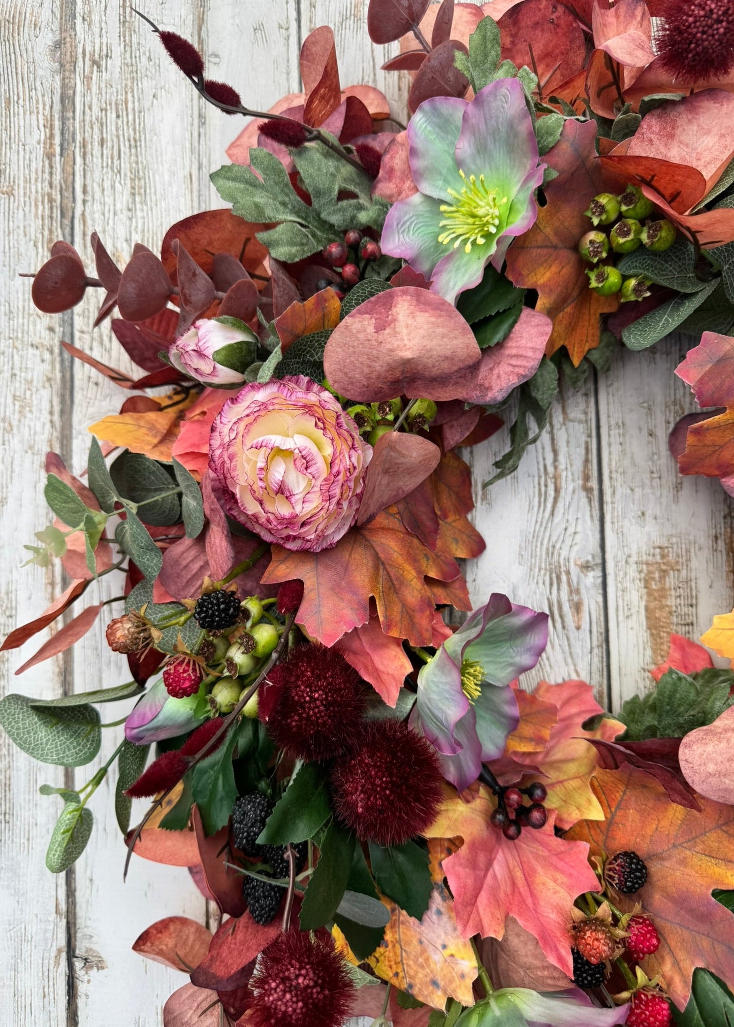 An autumn floral wreath with pink peonies, hollyhocks, burgundy berries, and fall leaves, arranged on a distressed white wooden background.