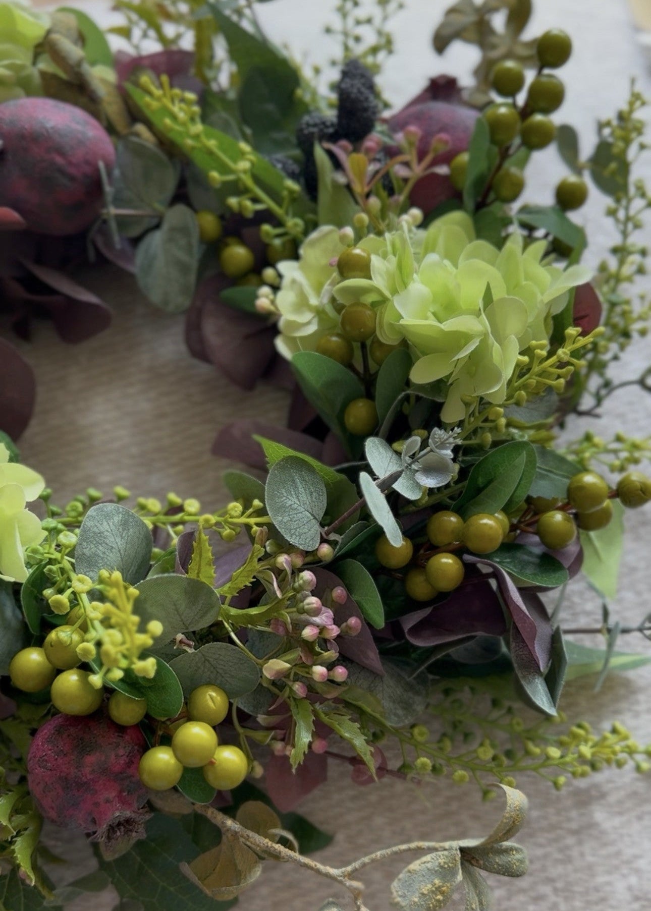 An artificial floral wreath featuring green hydrangea-like blooms, olive berries, eucalyptus, and muted burgundy leaves, arranged on a light beige fabric background.