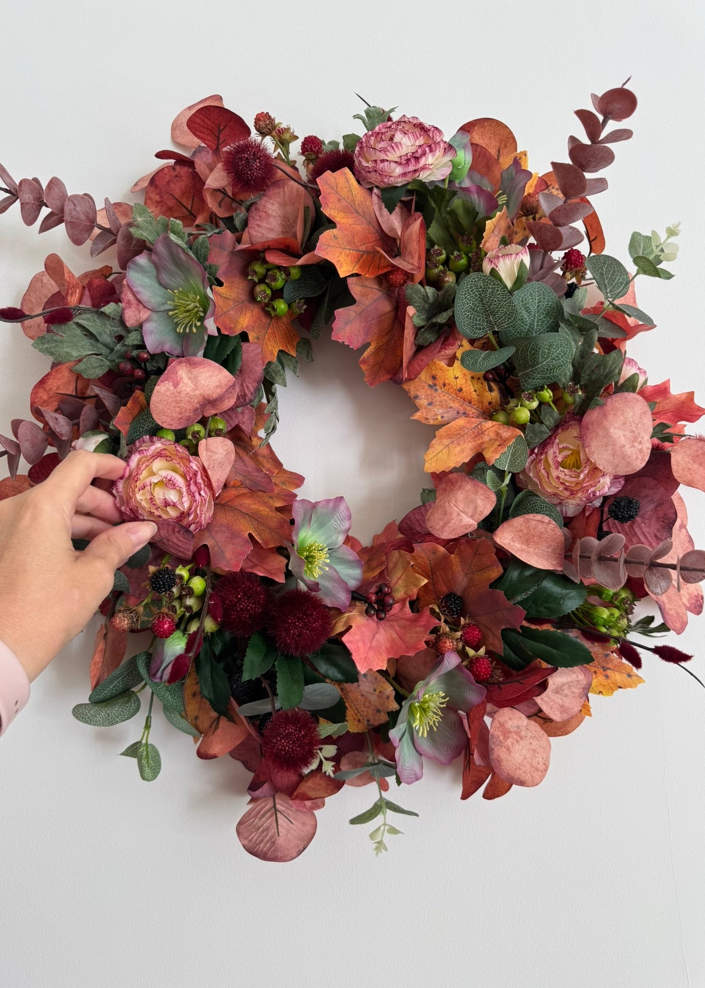 An autumn-themed floral wreath with pink roses, hollyhocks, eucalyptus, and fall leaves, shown against a plain white background with a hand adjusting one of the pink flowers on the left.