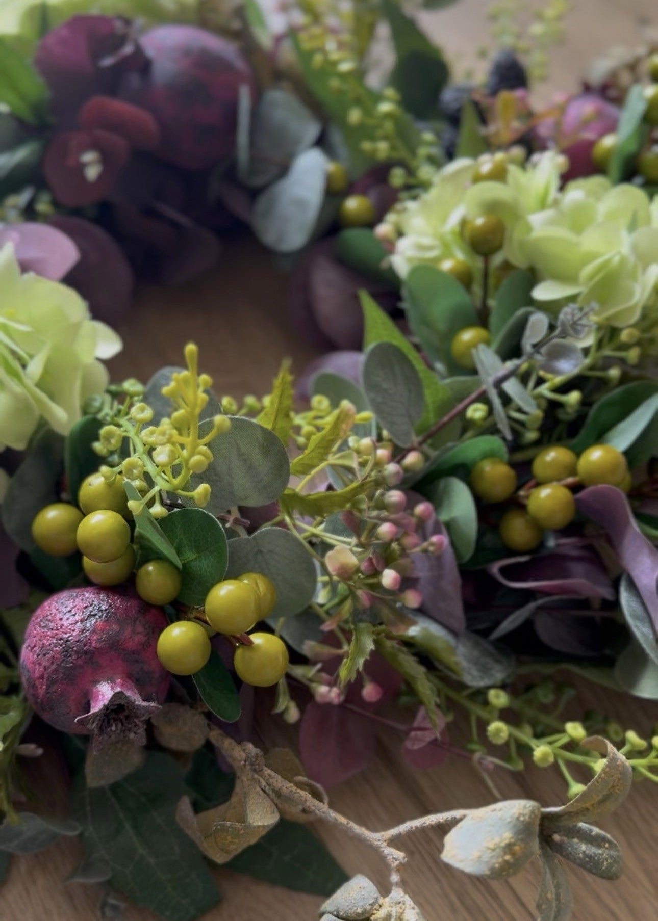 An artificial floral wreath with green eucalyptus leaves, yellow berries, purple hydrangeas, and pomegranate accents, displayed on a light wood floor.