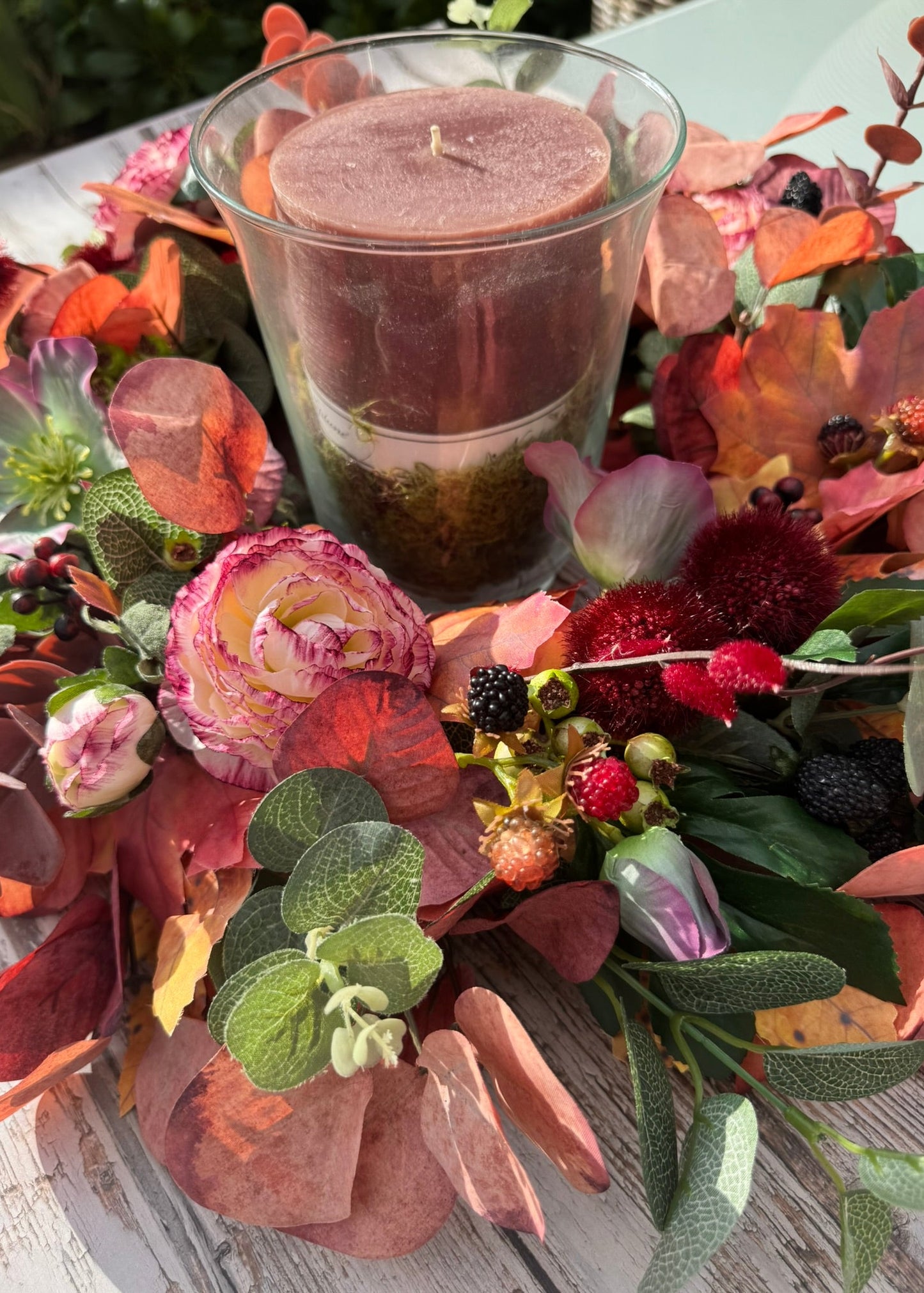 A layered candle in a clear glass holder sits surrounded by an autumn floral arrangement with pink roses, eucalyptus leaves, berries, and warm-toned foliage on a white wooden surface.