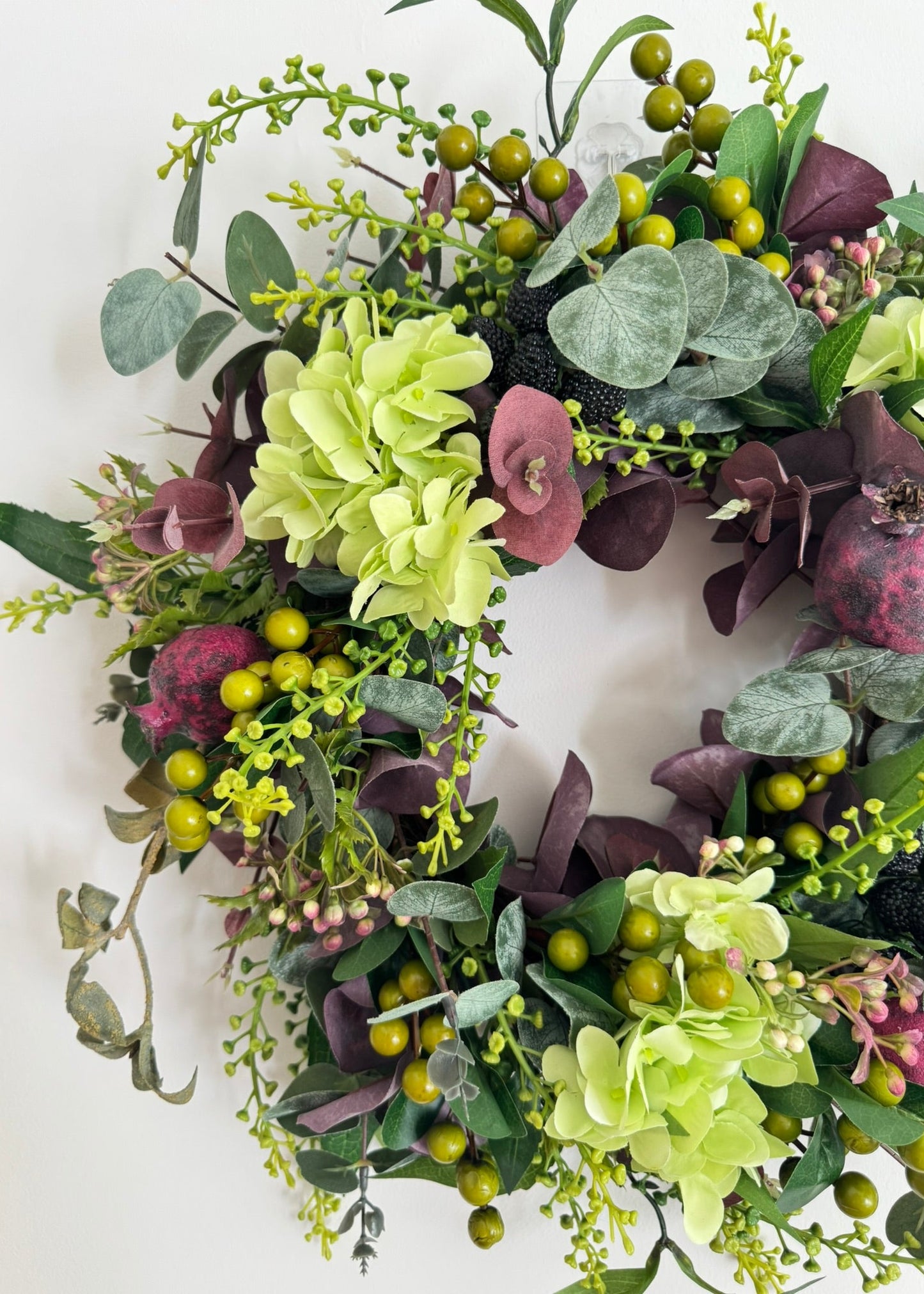An artificial floral wreath with light green hydrangeas, dusty pink flowers, eucalyptus leaves, dark purple foliage, and green berries, photographed against a plain white background.