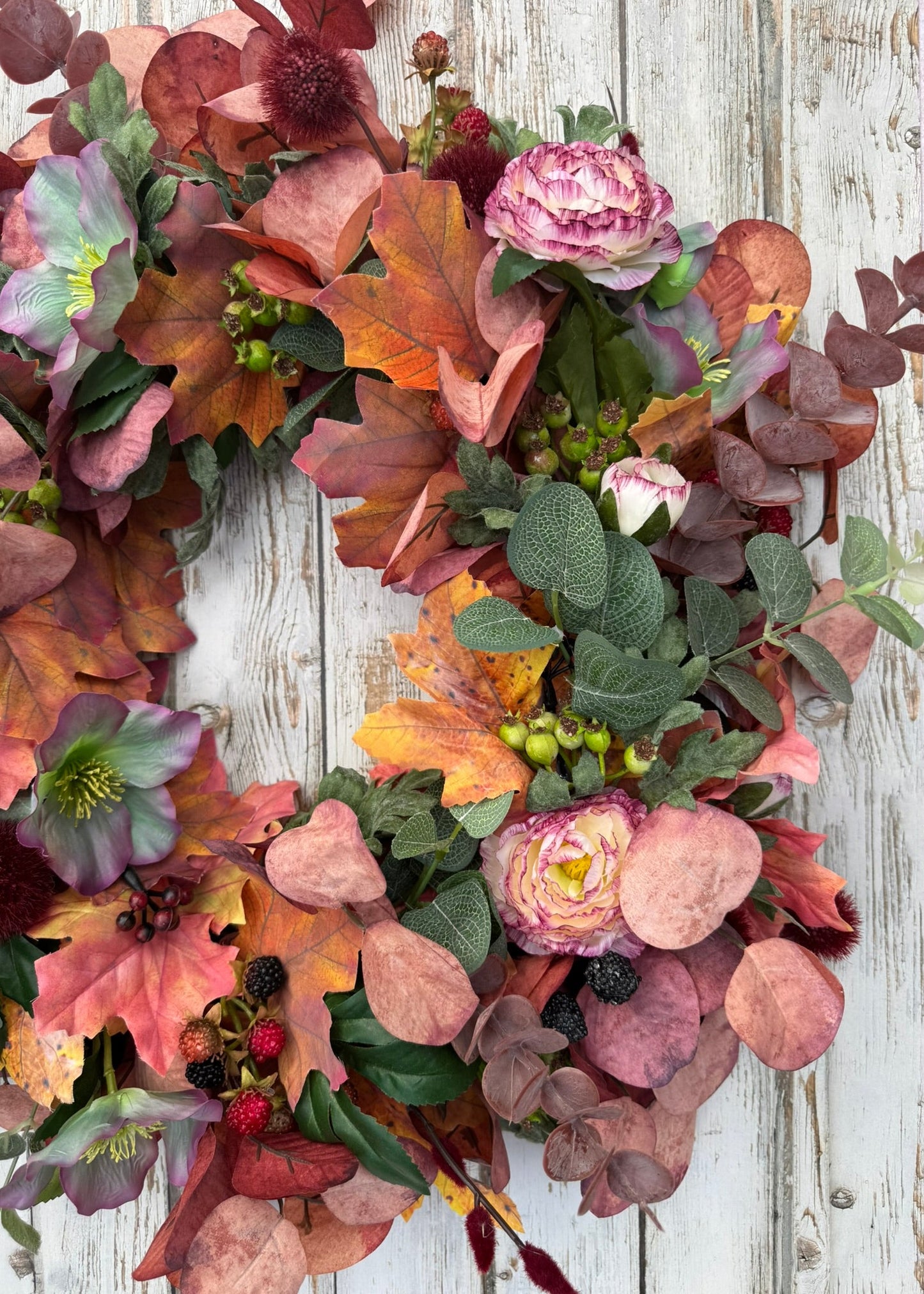An autumn floral wreath with pink roses, hollyhocks, eucalyptus, and fall leaves, displayed on a distressed white wood background.