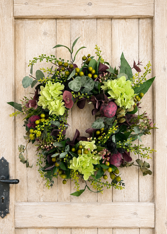 An artificial floral wreath with light green hydrangea-like blooms, dark burgundy berries, and green foliage, hanging on a light wood paneled door.