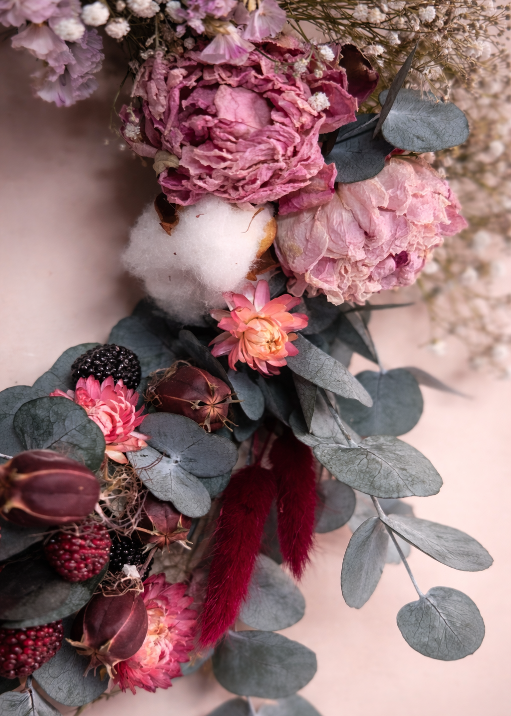 Close-up of a floral arrangement with pink and purple flowers, cotton, and green leaves on a soft pink background.