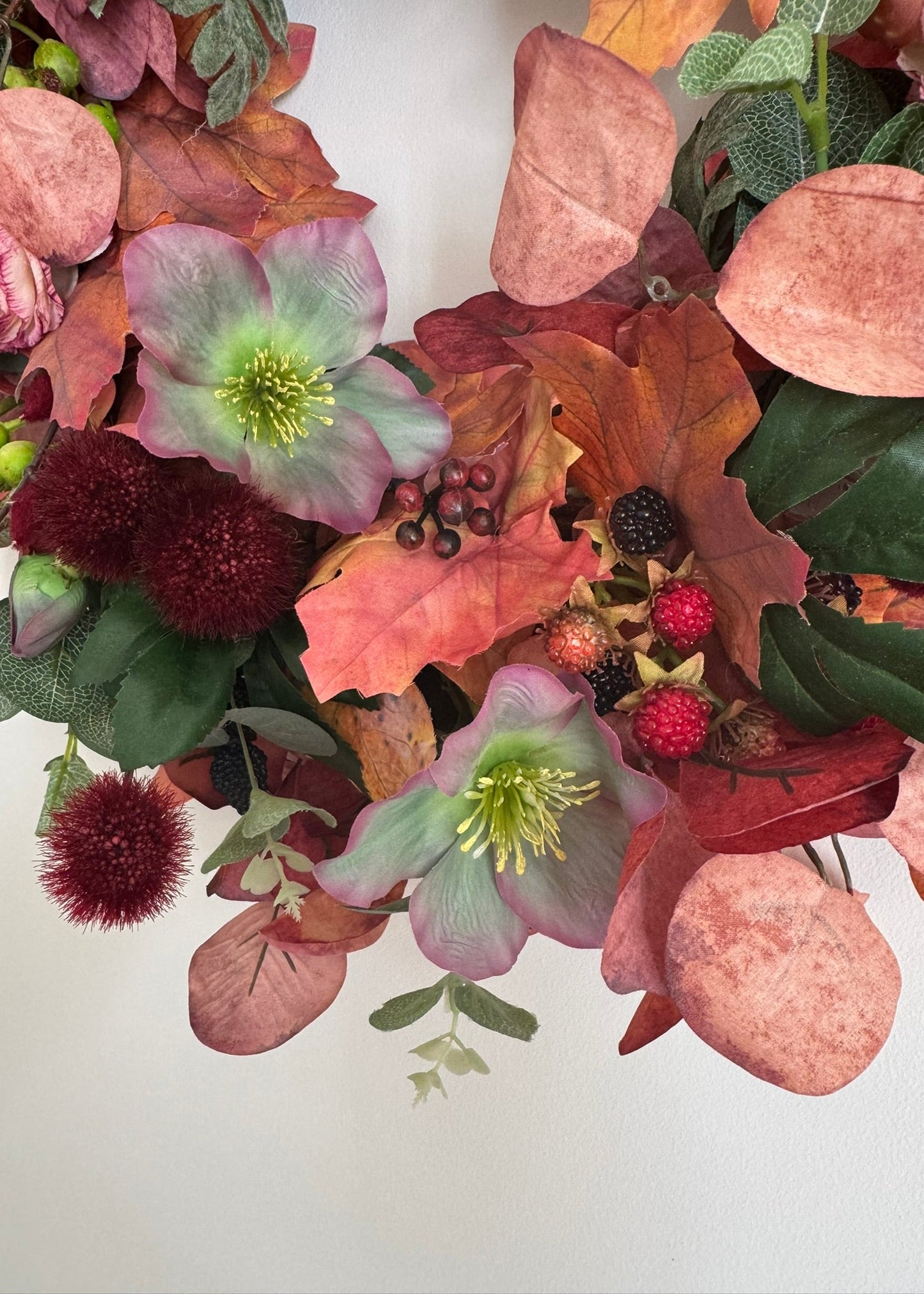 An artificial autumn floral wreath featuring pink hollyhocks, burgundy pom-pom flowers, and warm-toned fall leaves on a plain white background.