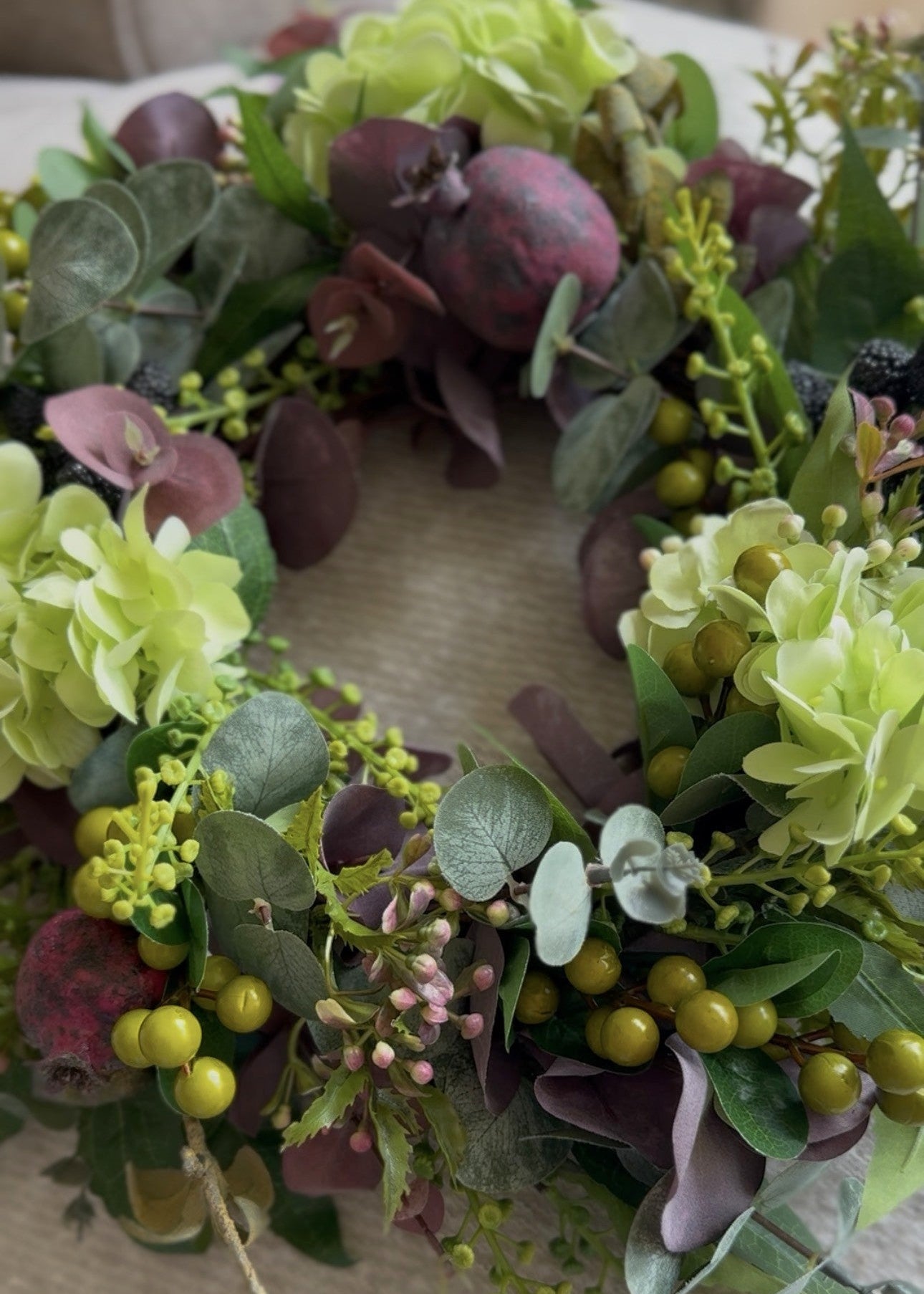 A lush artificial floral wreath featuring light green hydrangea-like blooms, dusty purple flowers, eucalyptus leaves, and small olive-green berries, arranged on a neutral fabric surface.