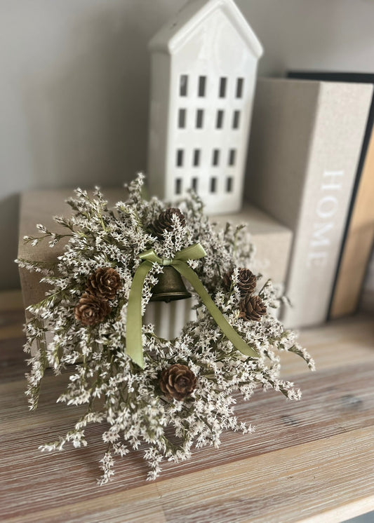 Decorative wreath with pinecones and a bell on a wooden surface with books in the background.