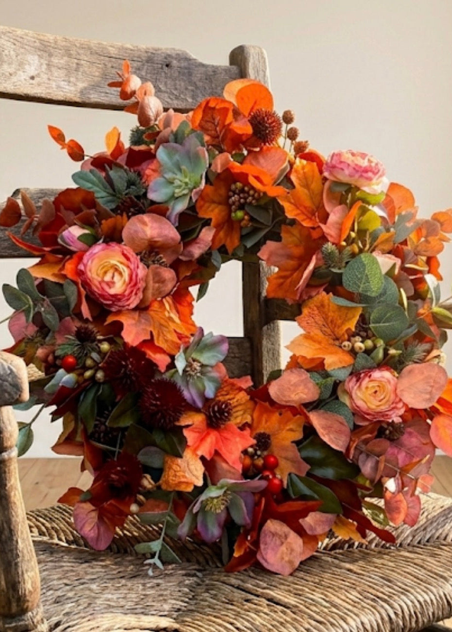 An autumn floral wreath with orange, red, pink, and green faux flowers and leaves, displayed on a rustic wooden chair against a plain beige wall.