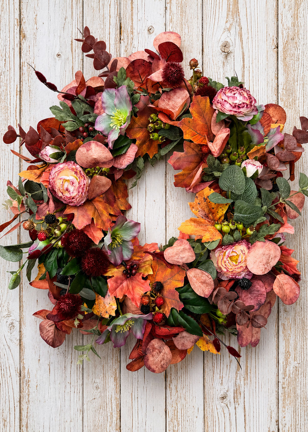 An autumn floral wreath with pink roses, orange and red leaves, green foliage, and small berries, displayed against a whitewashed wood plank background.