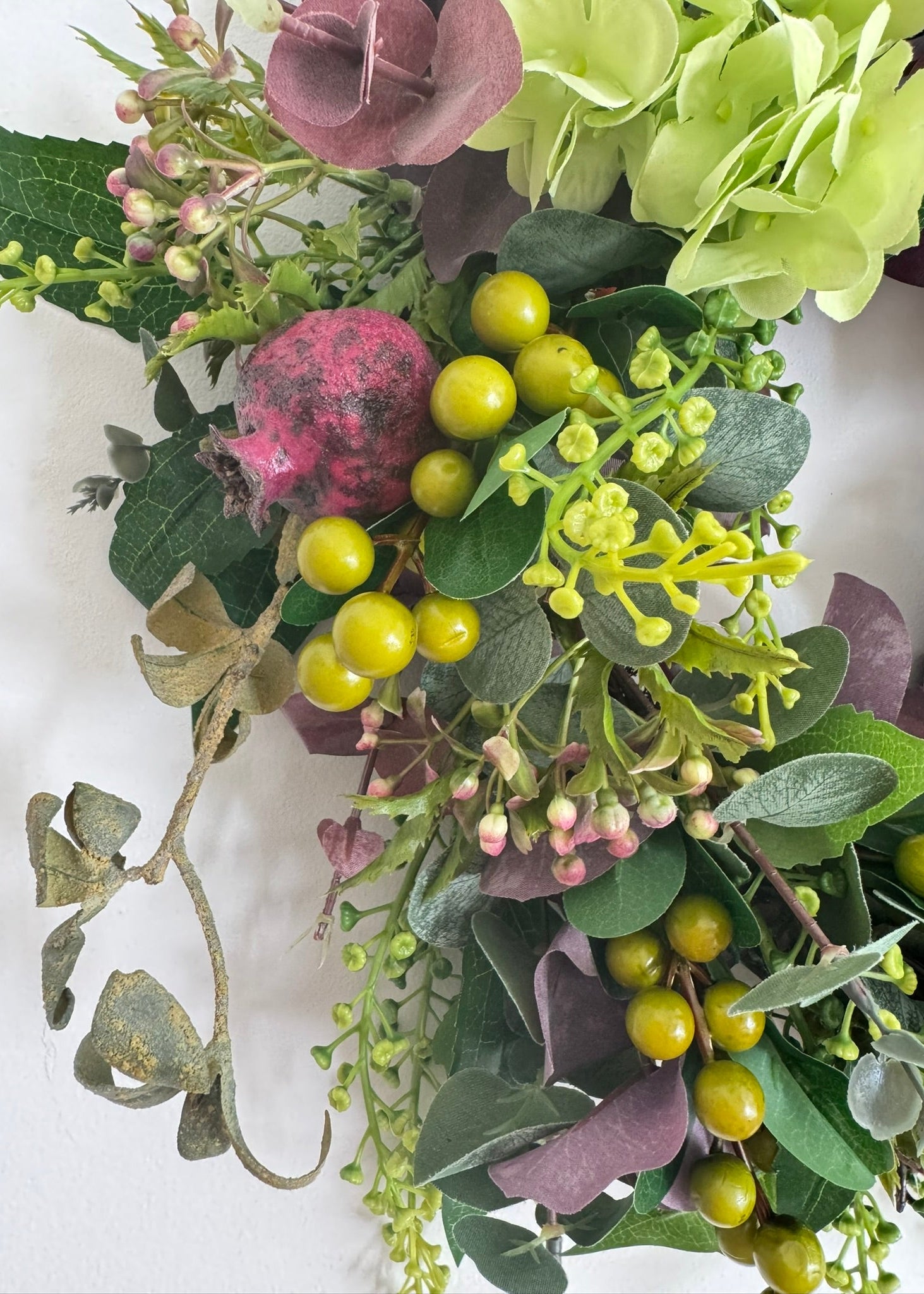 An artificial floral arrangement featuring green hydrangea blooms, yellow-green berries, a pink pomegranate, and green foliage on a white background.