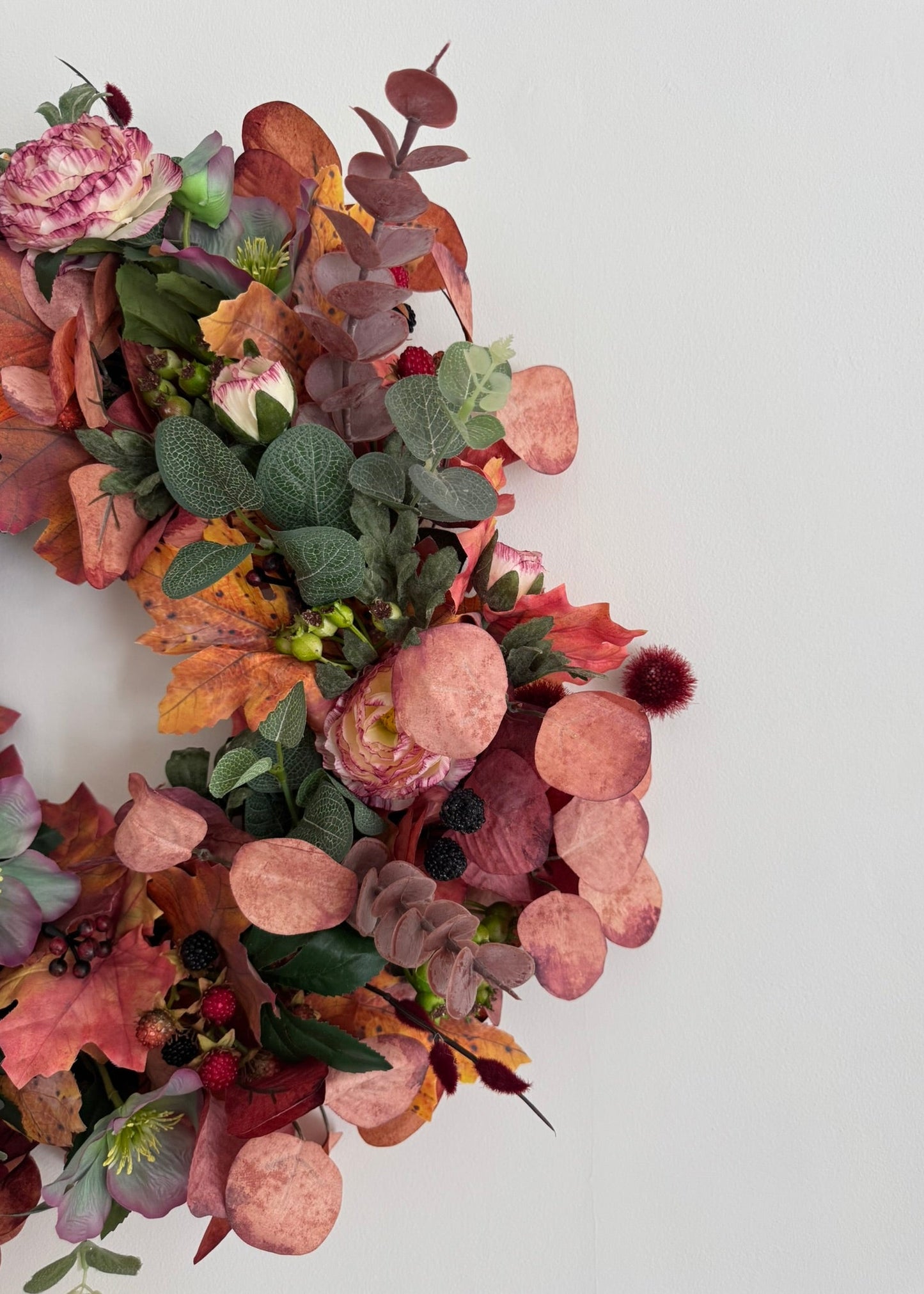 An autumn-themed floral wreath featuring pink ranunculus, dusty pink eucalyptus, orange and brown leaves, and green foliage, arranged on a plain white background.