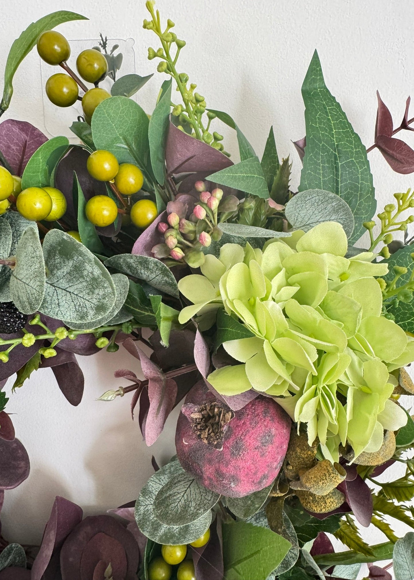 An artificial floral wreath featuring light green hydrangea-like blooms, sage green and dusty purple leaves, yellow berries, and a pink pomegranate accent, shown against a plain off-white wall.
