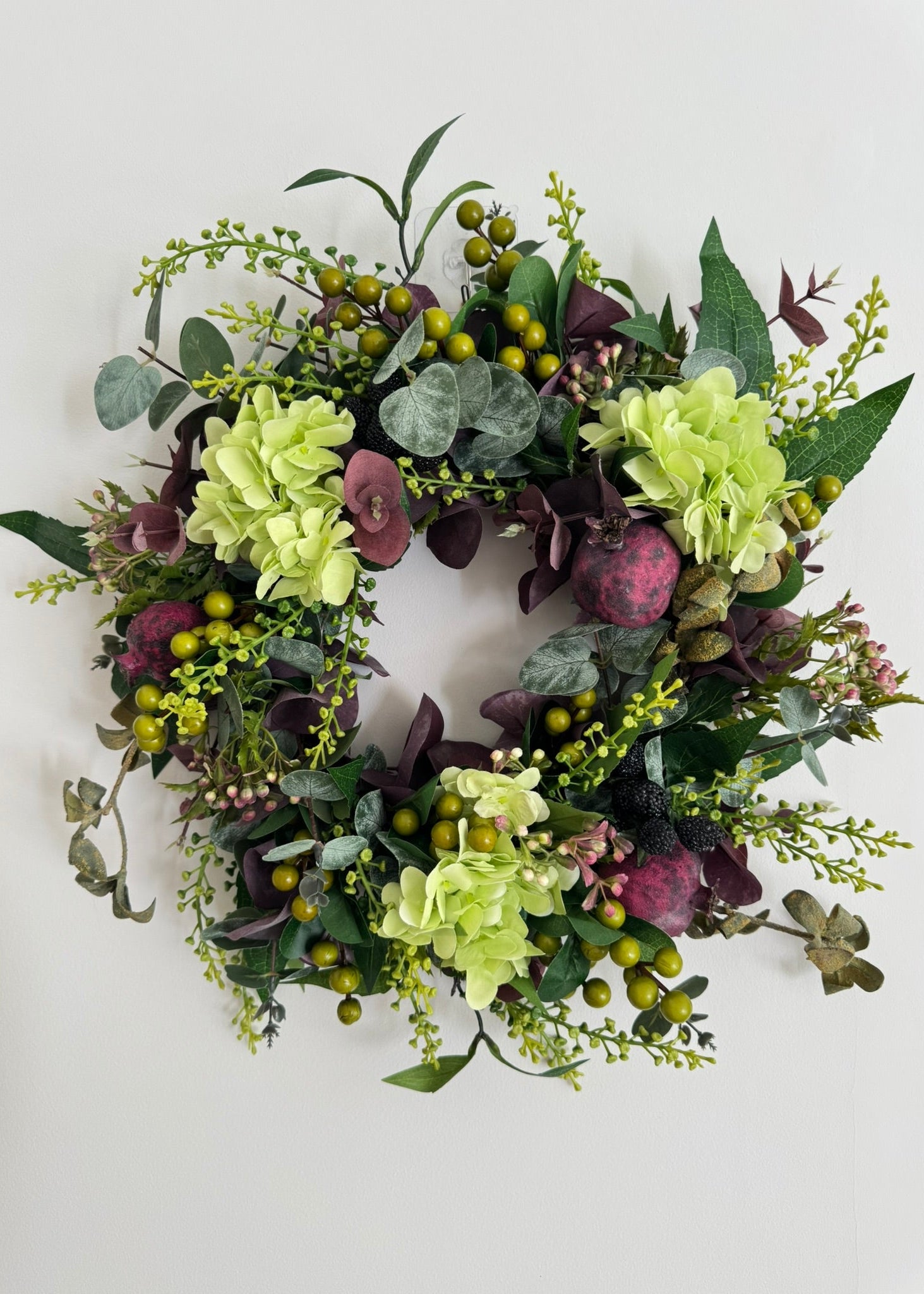 An artificial floral wreath featuring light green hydrangea-like blooms, dusty burgundy roses, sage green eucalyptus leaves, and small olive-green berries, displayed against a plain off-white background.