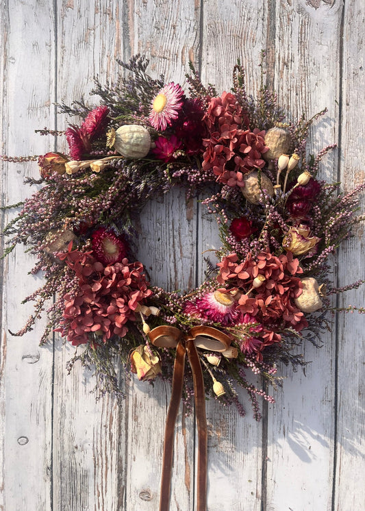 A dried floral wreath featuring a variety of autumnal flowers in shades of brown, burgundy, and pink, arranged with a brown velvet ribbon and designed to be displayed on a wall.