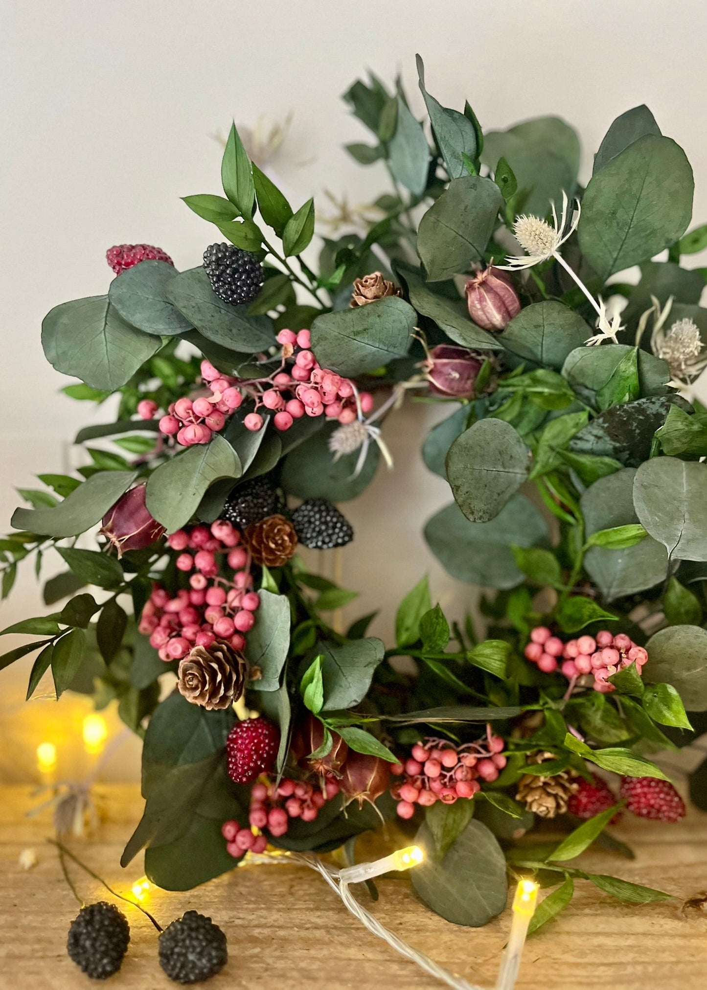 Decorative wreath with greenery, berries, and small pinecones on a wooden surface.