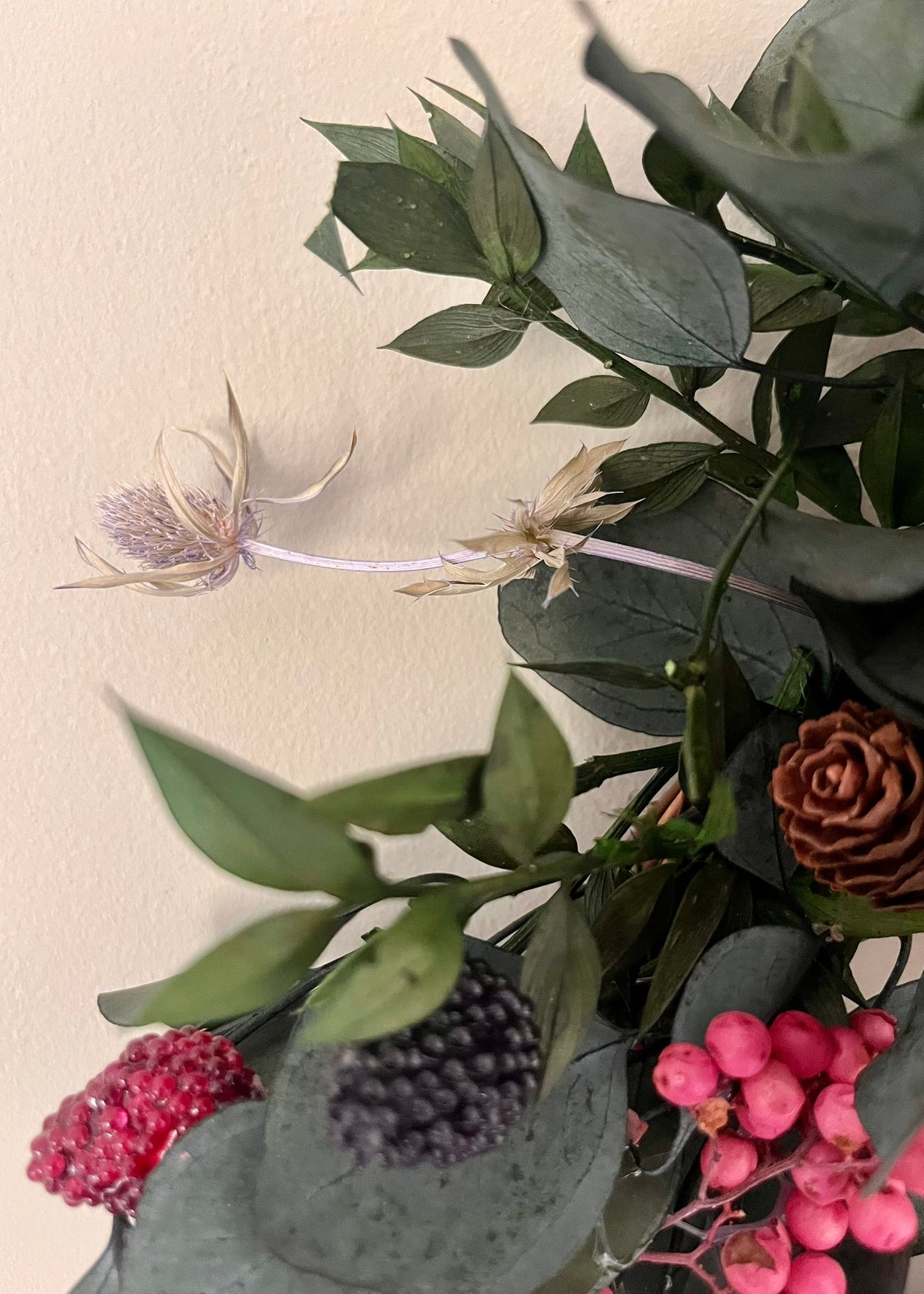 Close-up of a bouquet with green leaves, pink berries, and dried flowers on a beige background