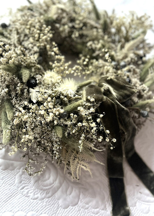A close-up of a handcrafted natural wreath with a combination of dried flowers and greenery, featuring an olive velvet ribbon bow.