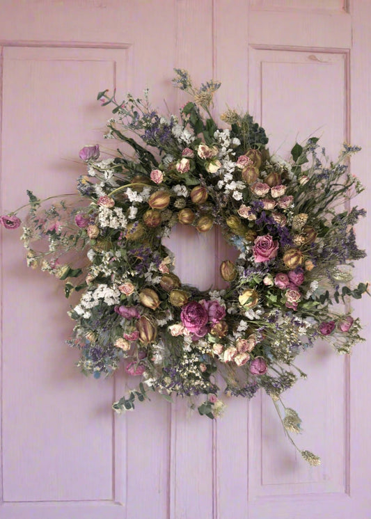 A dried floral wreath featuring a variety of flowers including pink ranunculas, eucalyptus, and other dried plants, arranged in a circular form against a pink background.