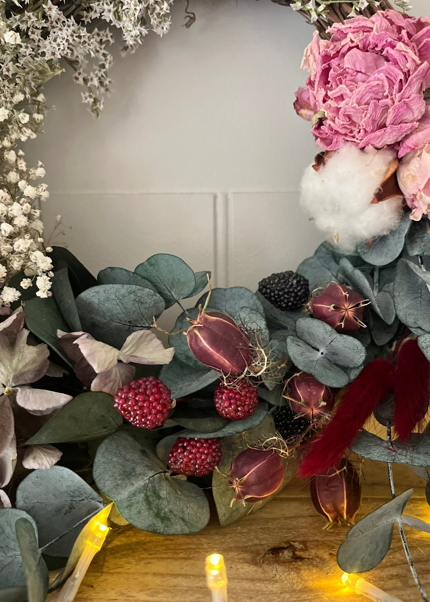 Floral wreath with pink flowers, green leaves, and berries on a wooden surface.