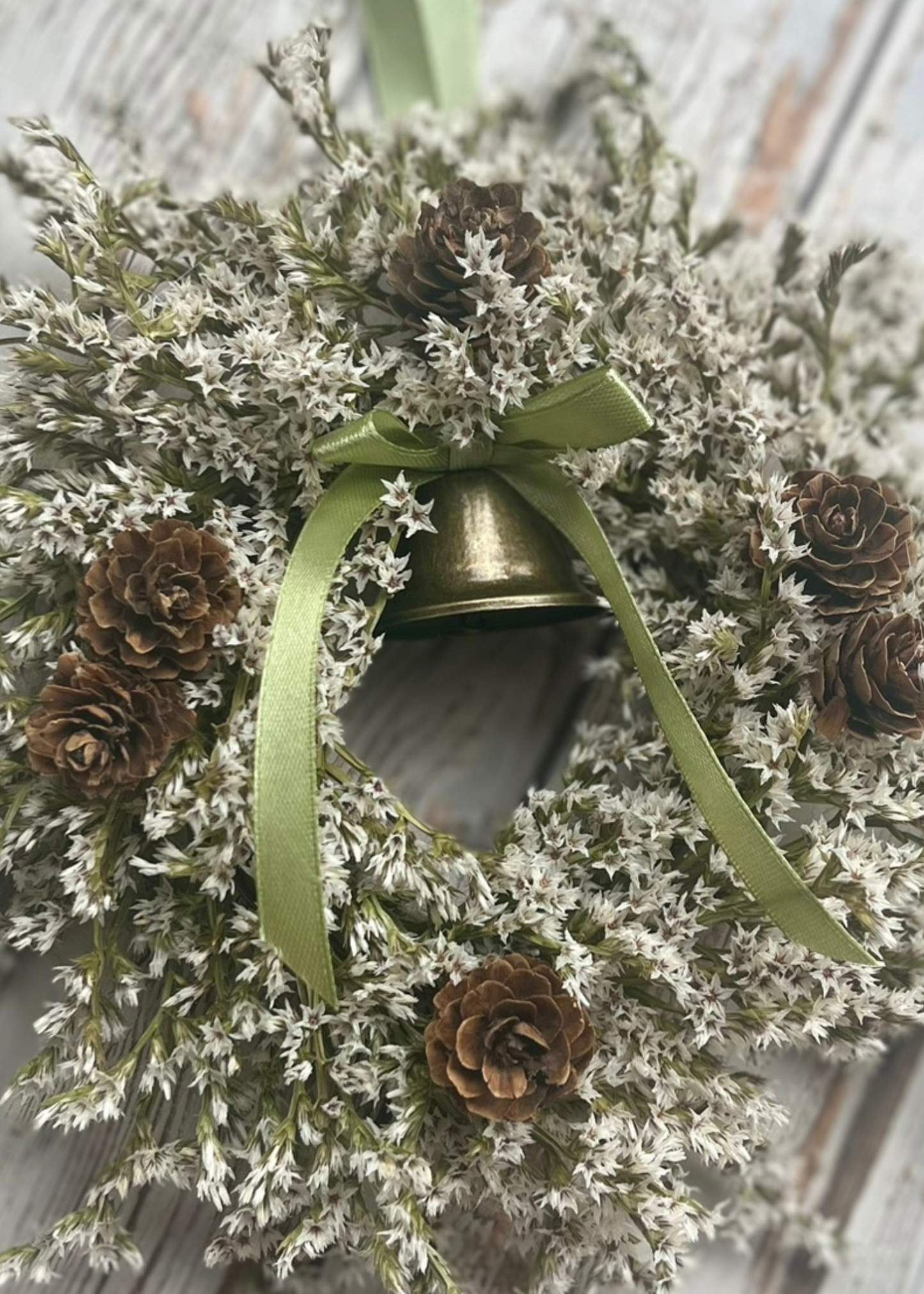 A mini Christmas wreath made of dried flowers including white statice, pine cones, and a brass bell, with satin olive or burgundy ribbon bow.