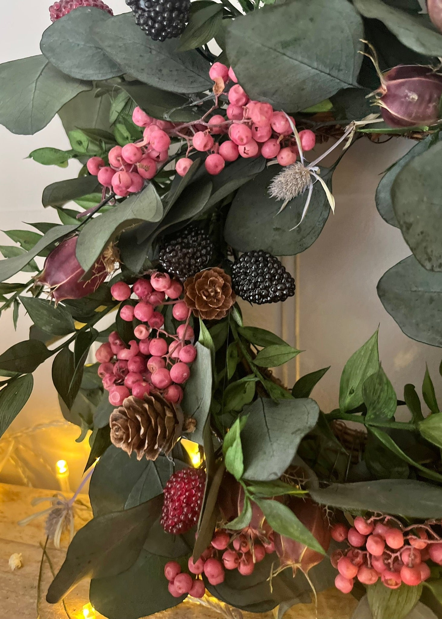 Decorative wreath with pink berries, green leaves, and pinecones on a neutral background.